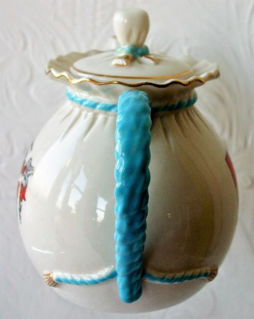 fig 1. An antique white porcelain teapot shaped like a tied bag, featuring a blue twisted rope handle, a ruffled collar, and a coat of arms for Torre Abbey on the body.