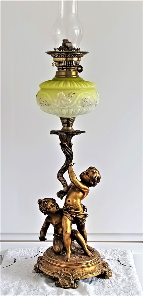 fig 1. An antique Victorian oil lamp featuring a gilded metal base with sculpted cherubs, a green glass reservoir, and a yellow ruffled glass shade with chimney.