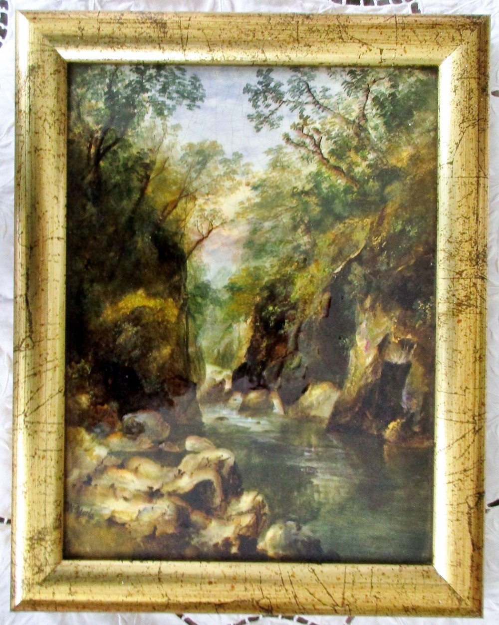 A framed rectangular ceramic plaque depicting a hand-painted river canyon landscape set within a distressed gold frame.