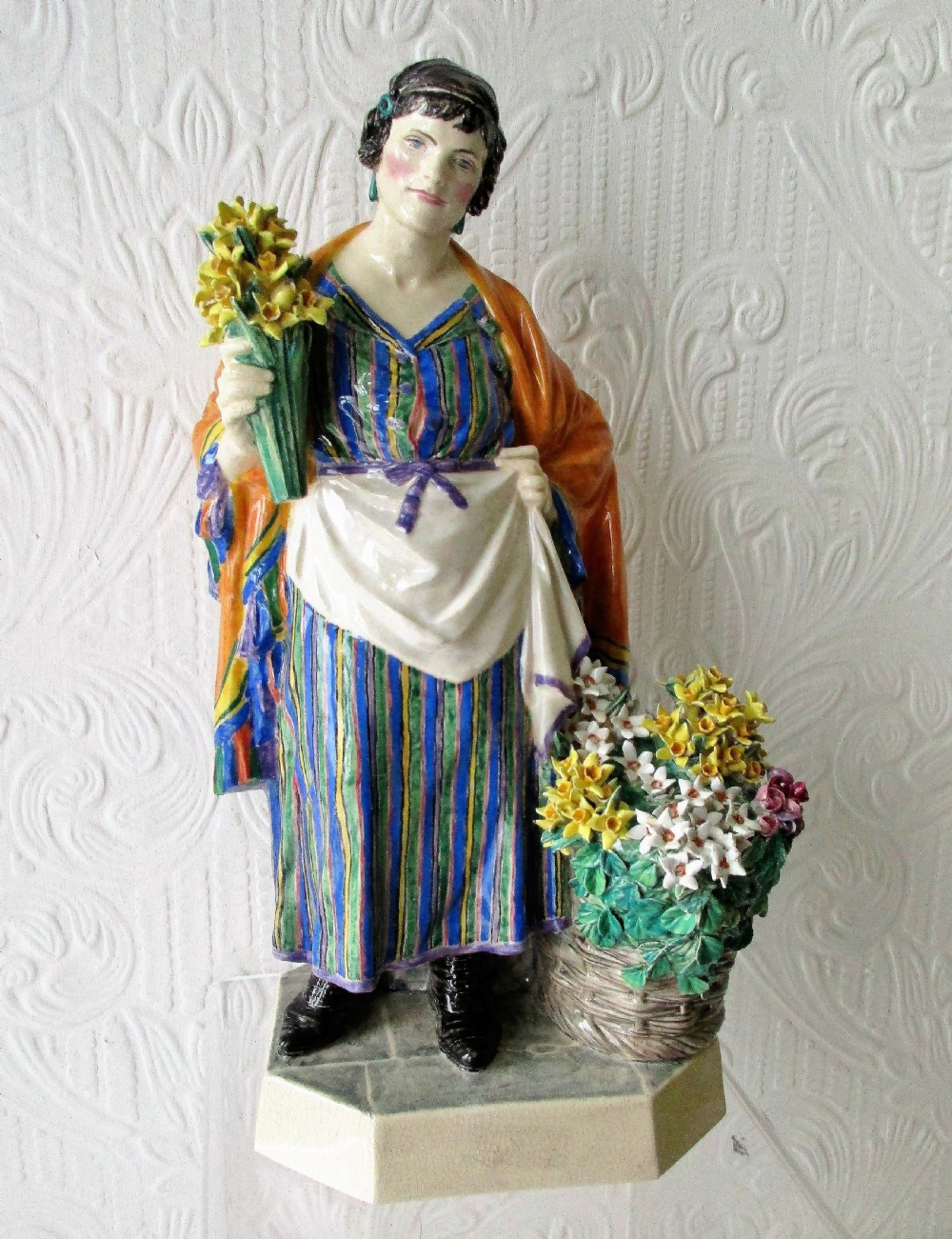 A glazed ceramic figurine depicting a female flower vendor holding a bunch of yellow daffodils next to a basket of flowers on an octagonal base.
