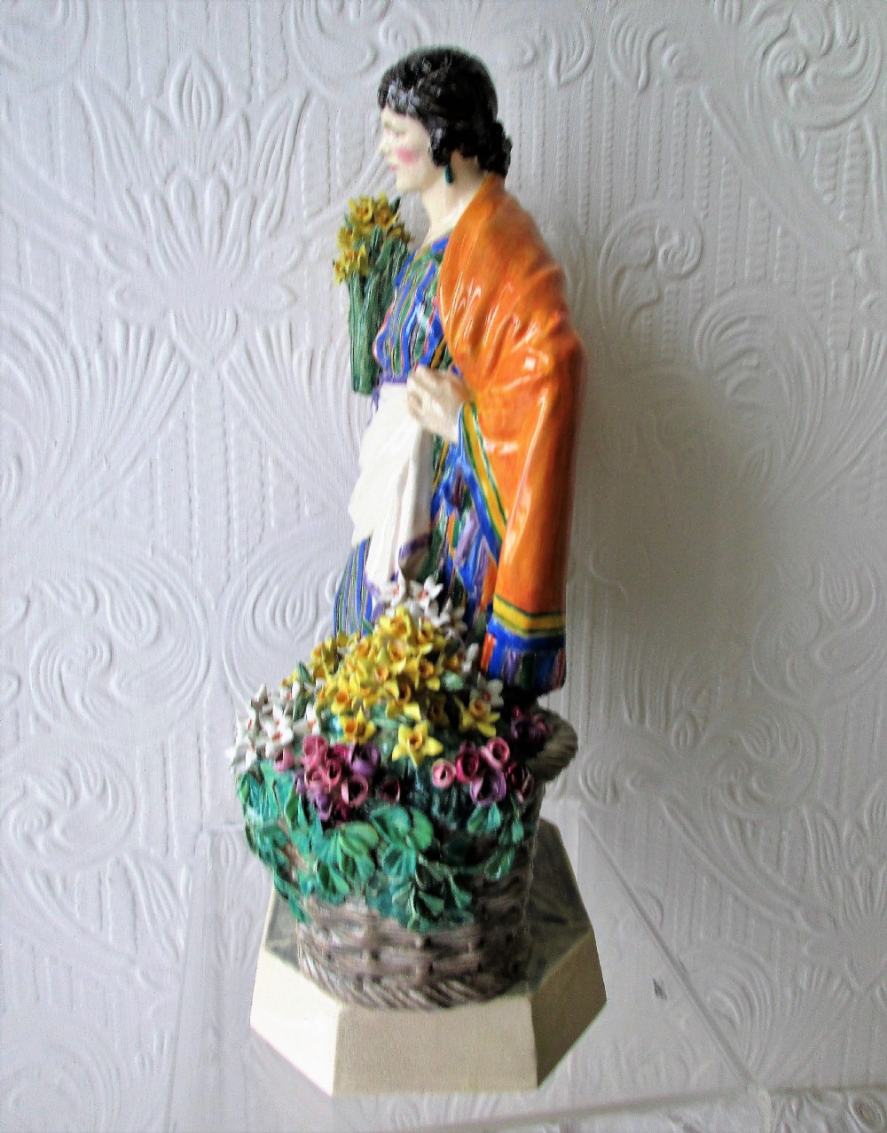 A glazed ceramic figurine depicting a female flower vendor holding a bunch of yellow daffodils next to a basket of flowers on an octagonal base. View II