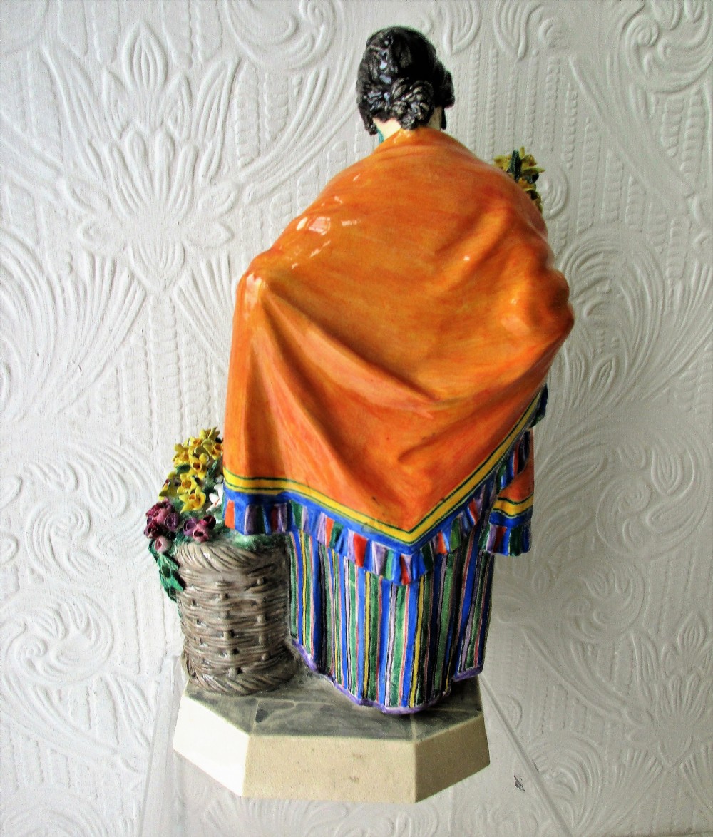 A glazed ceramic figurine depicting a female flower vendor holding a bunch of yellow daffodils next to a basket of flowers on an octagonal base. View III