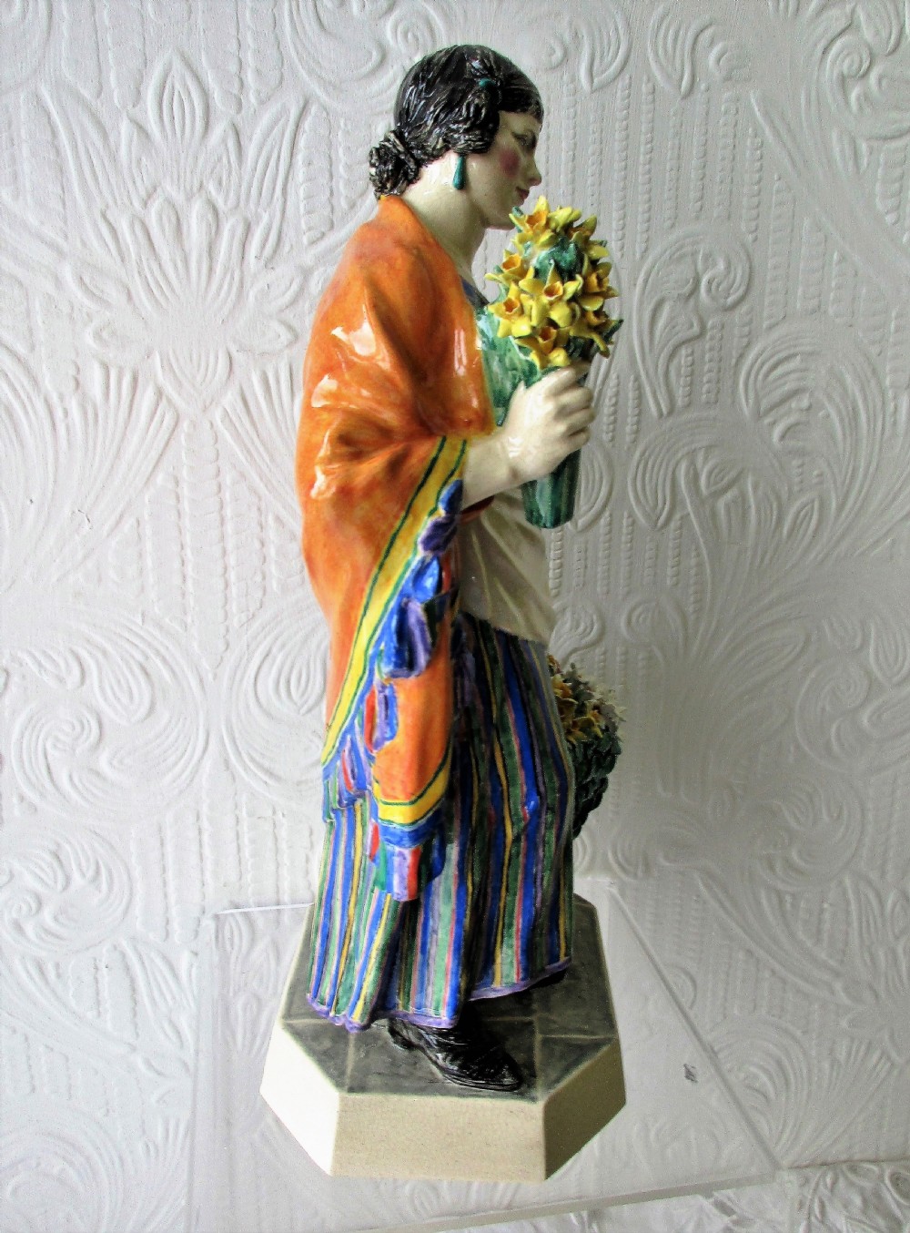 A glazed ceramic figurine depicting a female flower vendor holding a bunch of yellow daffodils next to a basket of flowers on an octagonal base. View IV