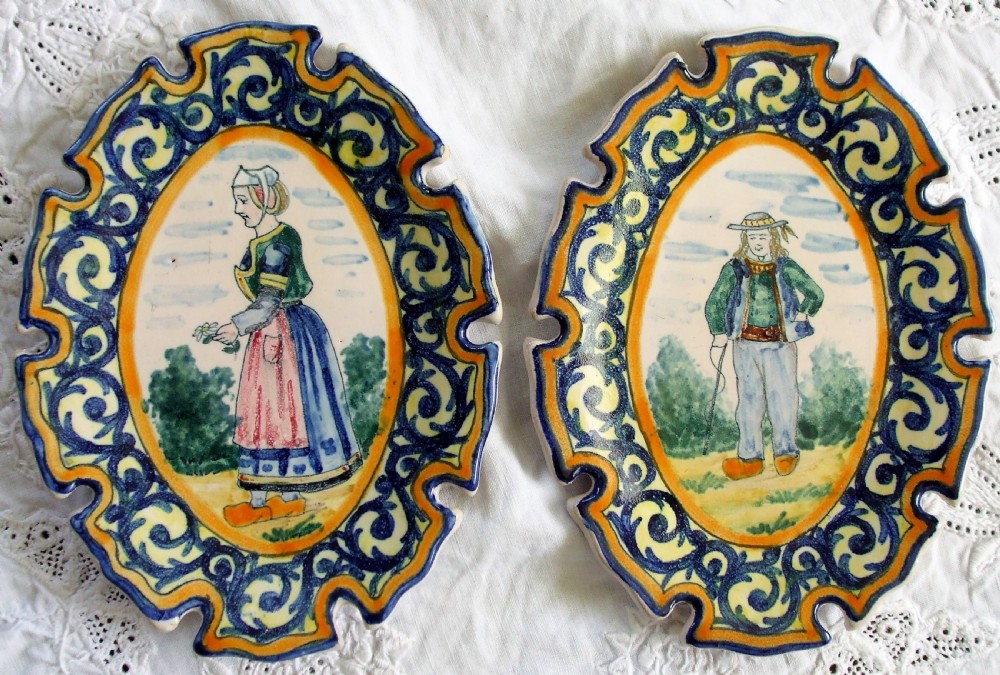 A pair of oval tin-glazed earthenware pin trays featuring hand-painted Breton figures surrounded by blue scrollwork and yellow borders with notched edges.