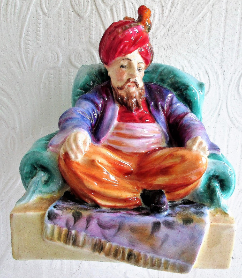 A glazed porcelain figurine depicting a bearded male figure wearing a red turban and seated cross-legged on a patterned carpet inside a green chair.