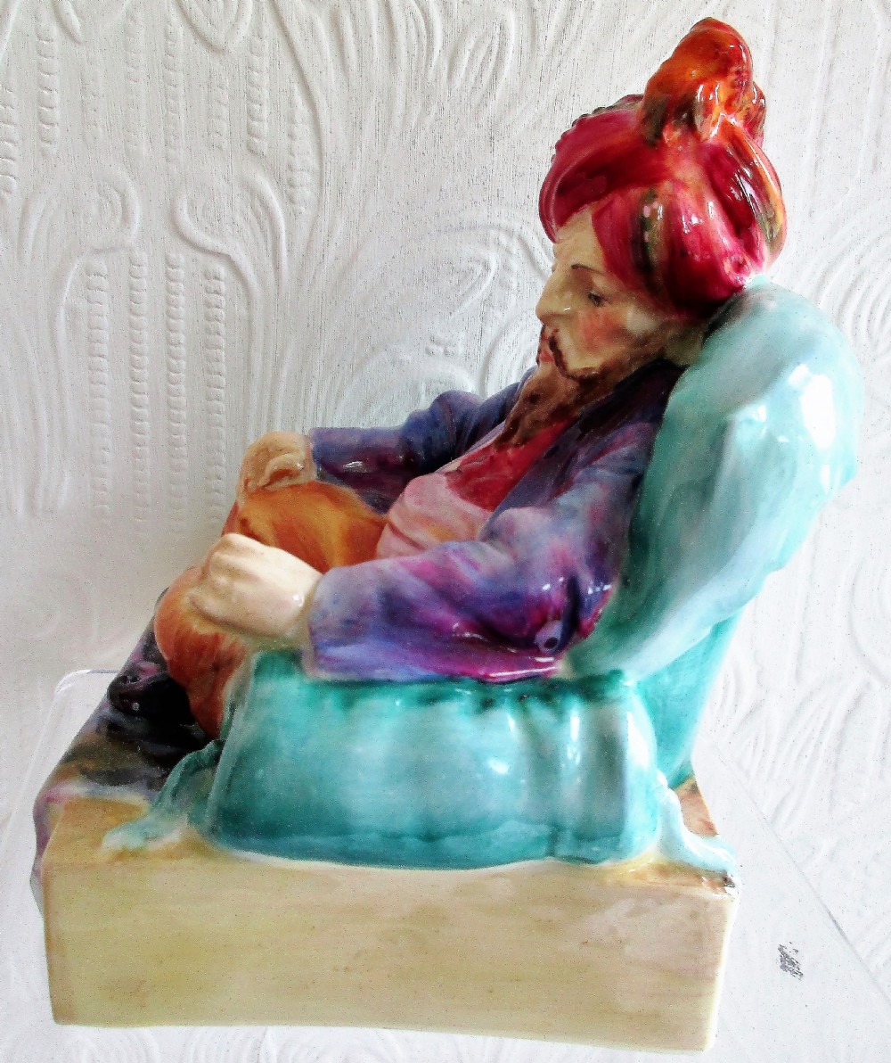 A glazed porcelain figurine depicting a bearded male figure wearing a red turban and seated cross-legged on a patterned carpet inside a green chair. View II