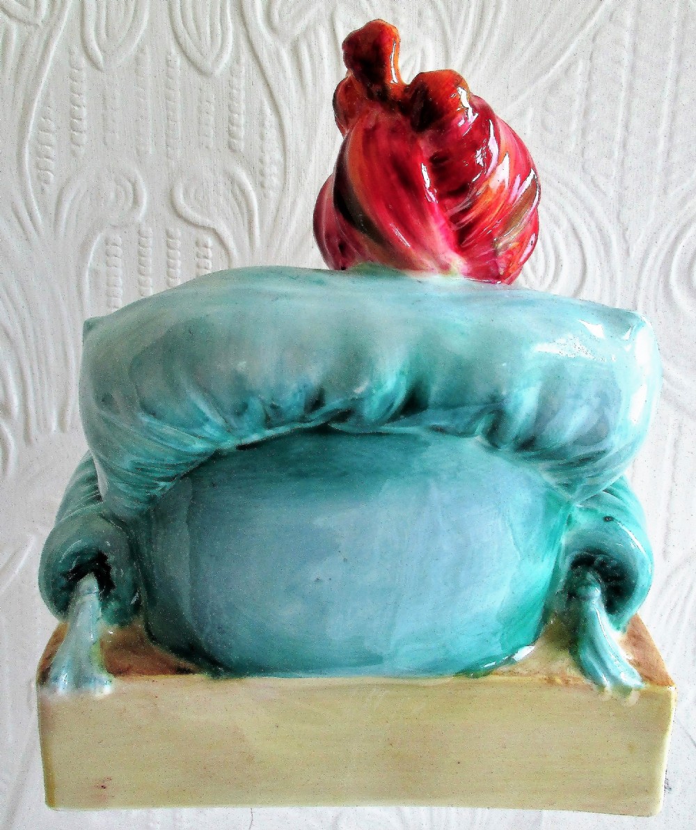 A glazed porcelain figurine depicting a bearded male figure wearing a red turban and seated cross-legged on a patterned carpet inside a green chair. View III