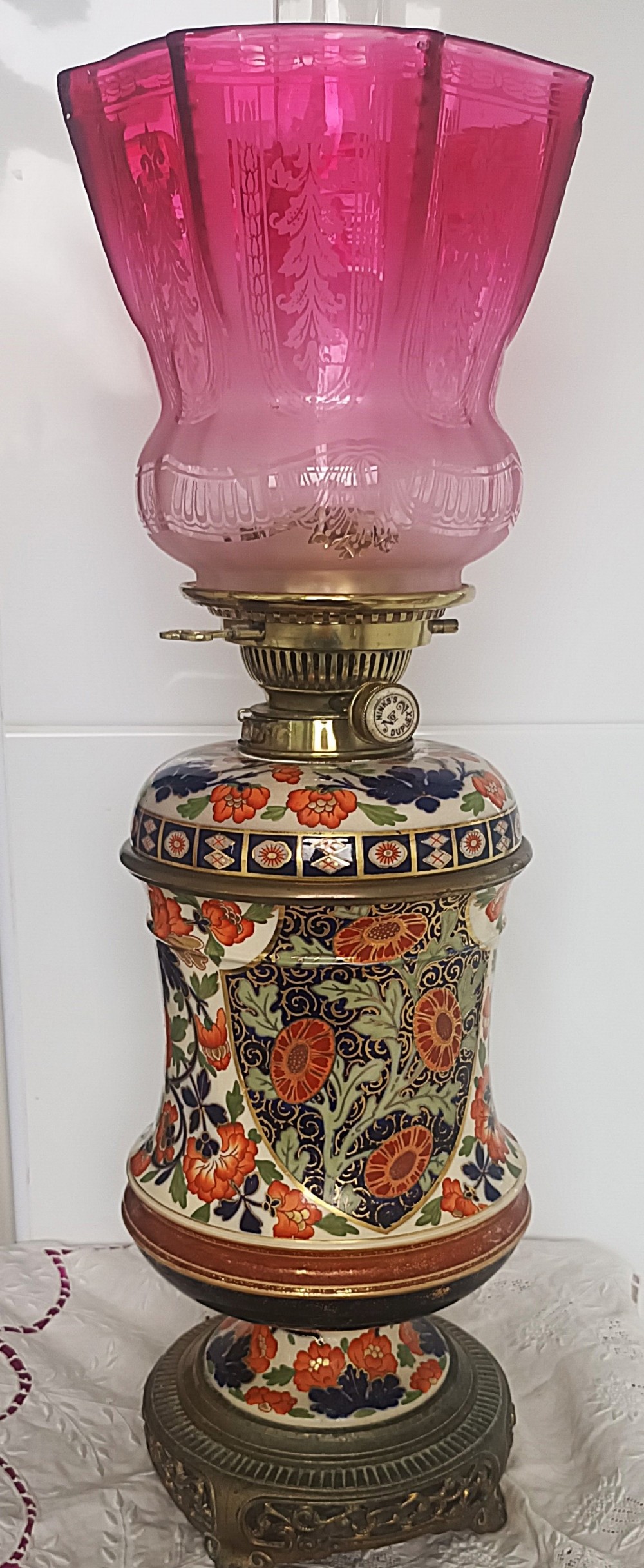 A Victorian ceramic oil lamp featuring an etched pink glass shade, brass Duplex burner mechanism, and hand-painted floral ceramic body on a metal base.