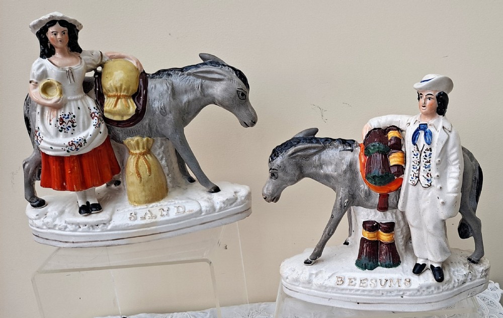 A pair of Victorian Staffordshire earthenware pottery figures depicting street traders named Sand and Beesums standing beside saddled grey donkeys.