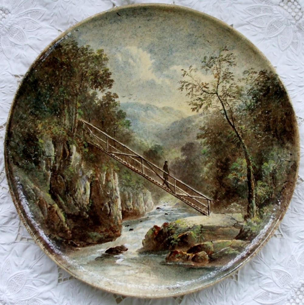 A circular Victorian ceramic plaque featuring a hand-painted landscape of a wooden footbridge crossing a river in North Wales.