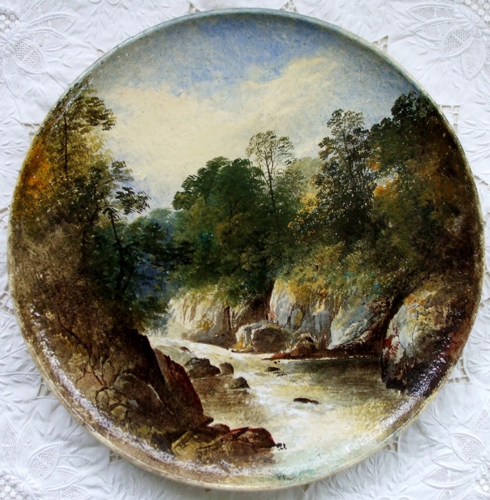 A circular Victorian earthenware plaque featuring a hand-painted landscape scene of a river running through a wooded gorge.