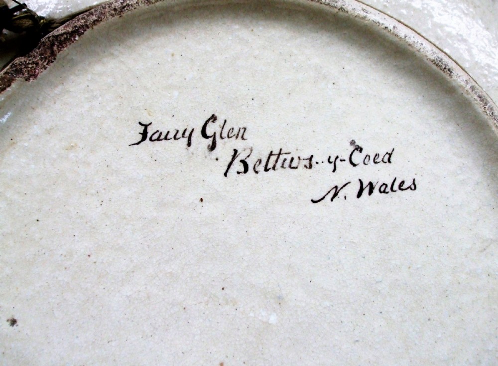 A circular Victorian earthenware plaque featuring a hand-painted landscape scene of a river running through a wooded gorge. View III