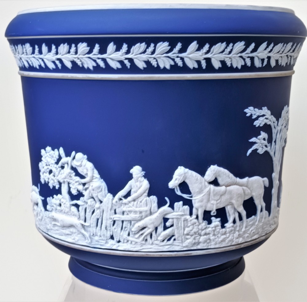 A cobalt blue ceramic jasperware jardiniere decorated with white raised relief scenes of a traditional hunt surrounded by foliage and a leaf band along the rim. View II