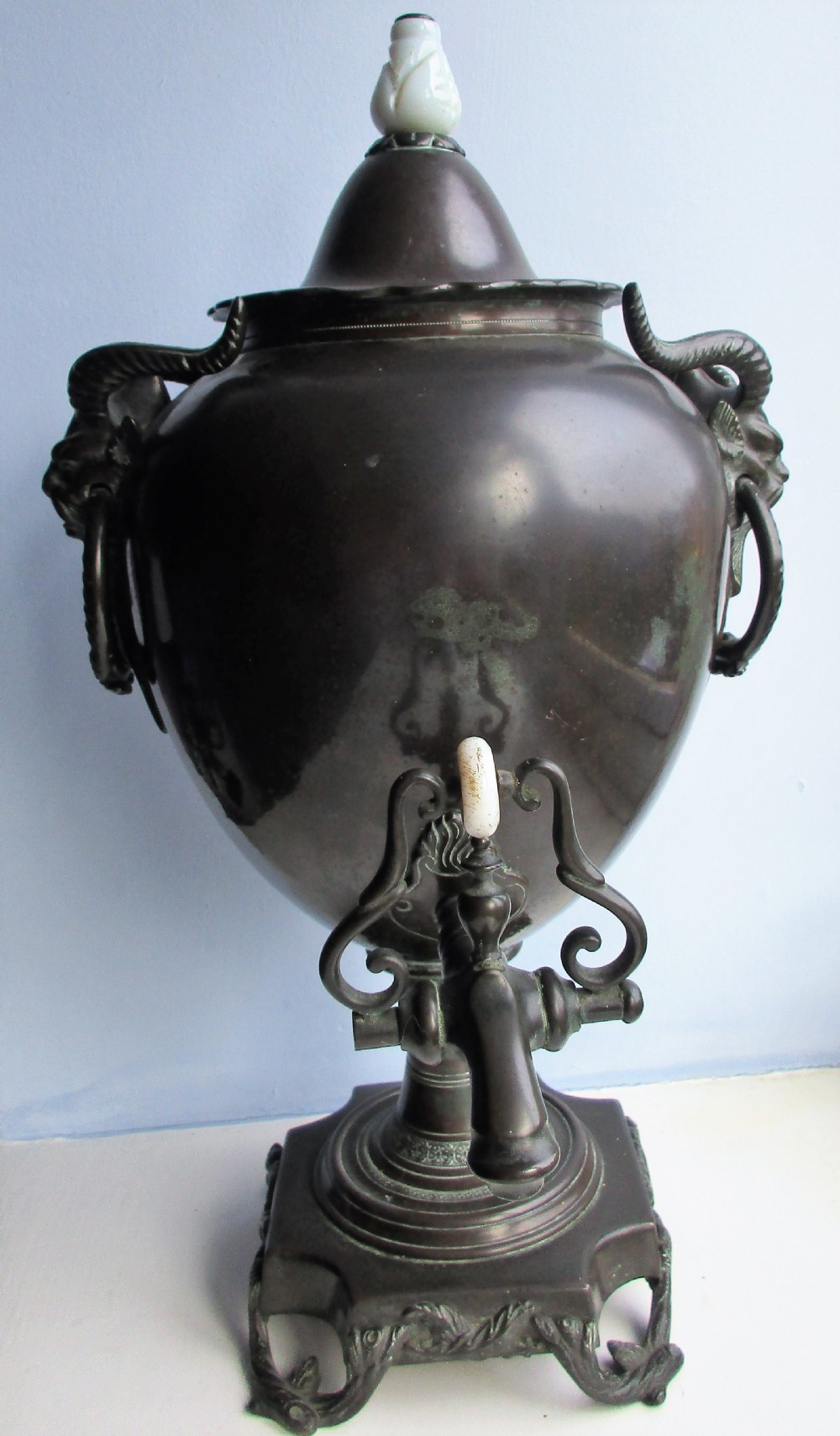 A dark patinated metal Victorian tea urn featuring a bulbous body, twin side handles, a front spigot with a white handle, and a pointed lid.