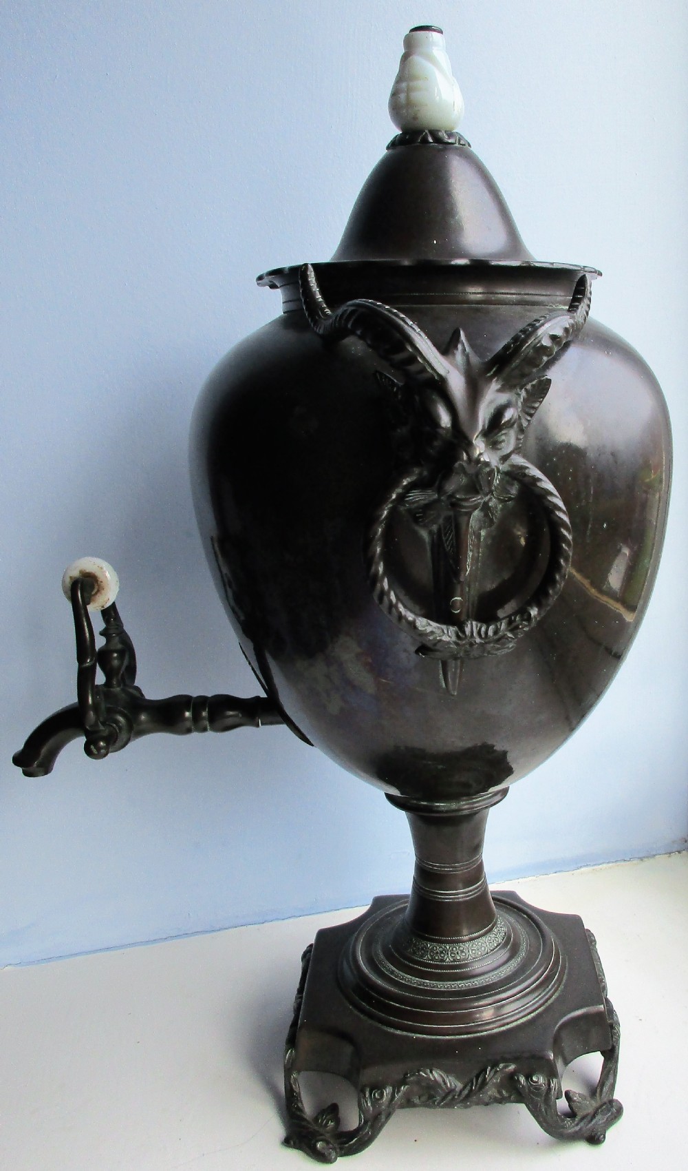 A dark patinated metal Victorian tea urn featuring a bulbous body, twin side handles, a front spigot with a white handle, and a pointed lid. View II
