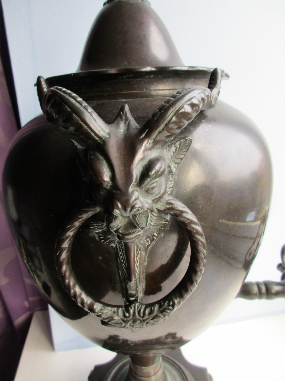 A dark patinated metal Victorian tea urn featuring a bulbous body, twin side handles, a front spigot with a white handle, and a pointed lid. View V