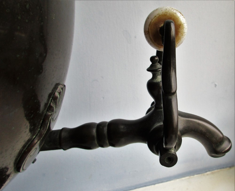 A dark patinated metal Victorian tea urn featuring a bulbous body, twin side handles, a front spigot with a white handle, and a pointed lid. View VI