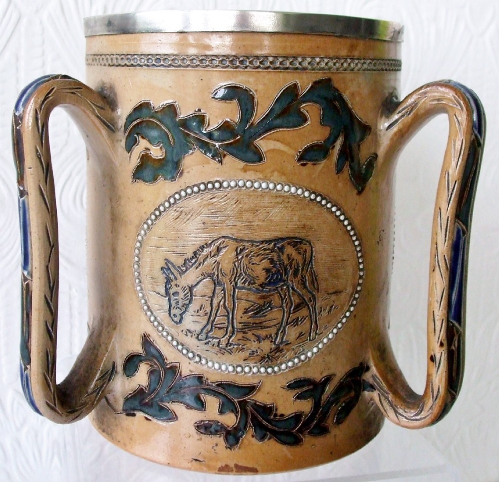 An antique tan stoneware tyg featuring three large handles, a silver-mounted rim, and an incised central medallion depicting a donkey surrounded by dark foliage scrollwork.