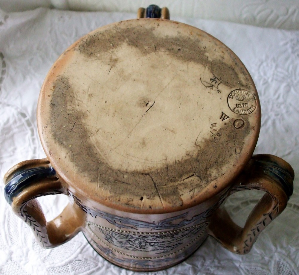 An antique tan stoneware tyg featuring three large handles, a silver-mounted rim, and an incised central medallion depicting a donkey surrounded by dark foliage scrollwork. View IV
