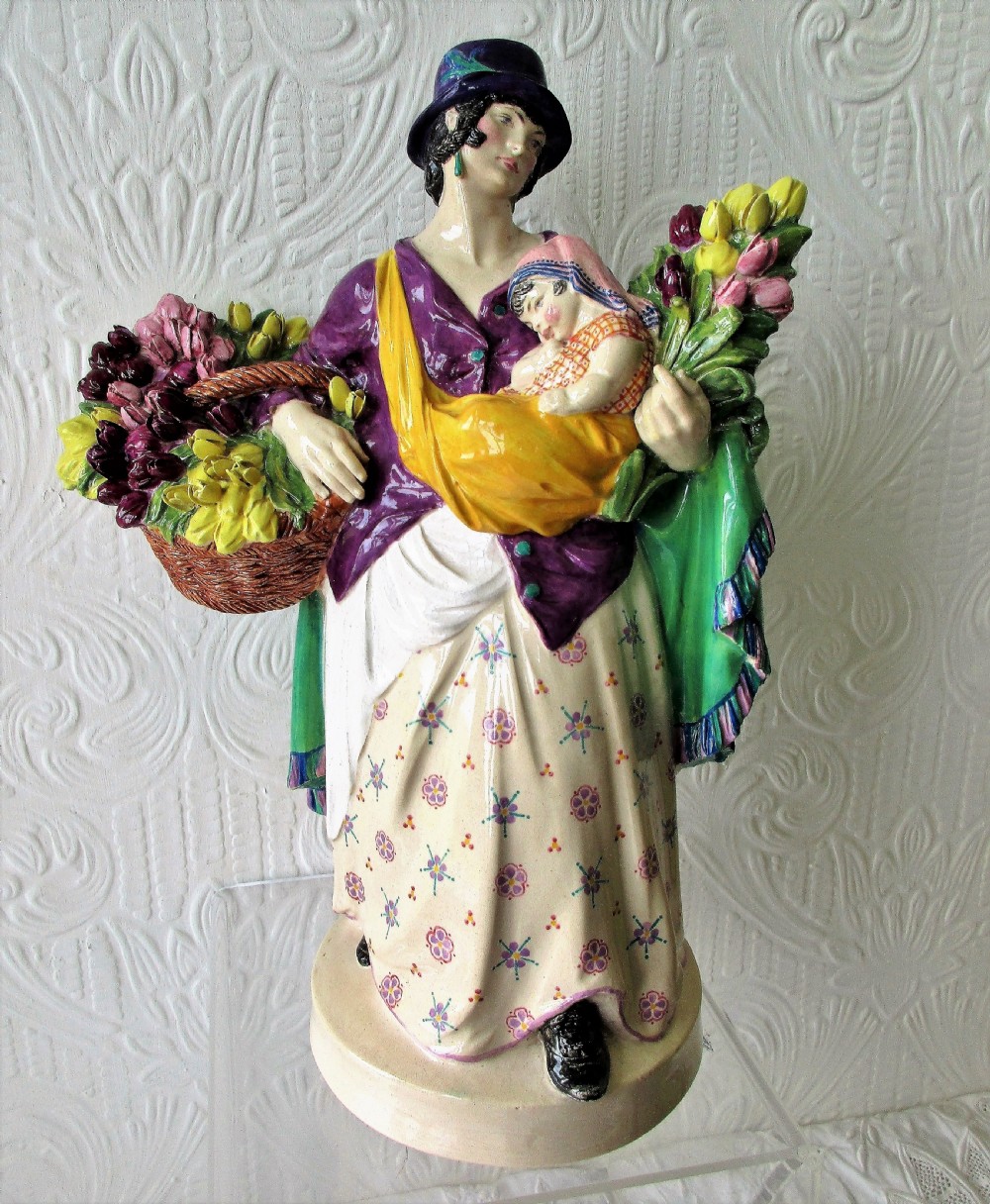 A polychrome glazed ceramic figurine depicting a woman wearing a purple jacket and blue hat, carrying a baby in a yellow sling and a basket of colourful flowers.