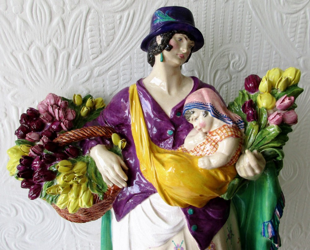 A polychrome glazed ceramic figurine depicting a woman wearing a purple jacket and blue hat, carrying a baby in a yellow sling and a basket of colourful flowers. View II