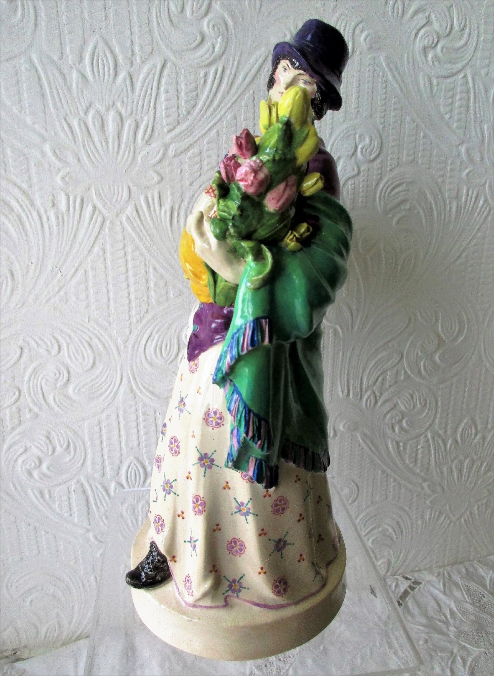 A polychrome glazed ceramic figurine depicting a woman wearing a purple jacket and blue hat, carrying a baby in a yellow sling and a basket of colourful flowers. View III