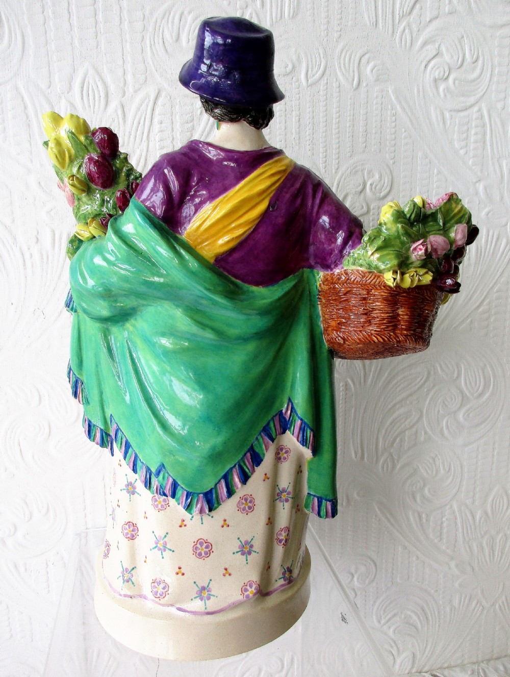 A polychrome glazed ceramic figurine depicting a woman wearing a purple jacket and blue hat, carrying a baby in a yellow sling and a basket of colourful flowers. View IV
