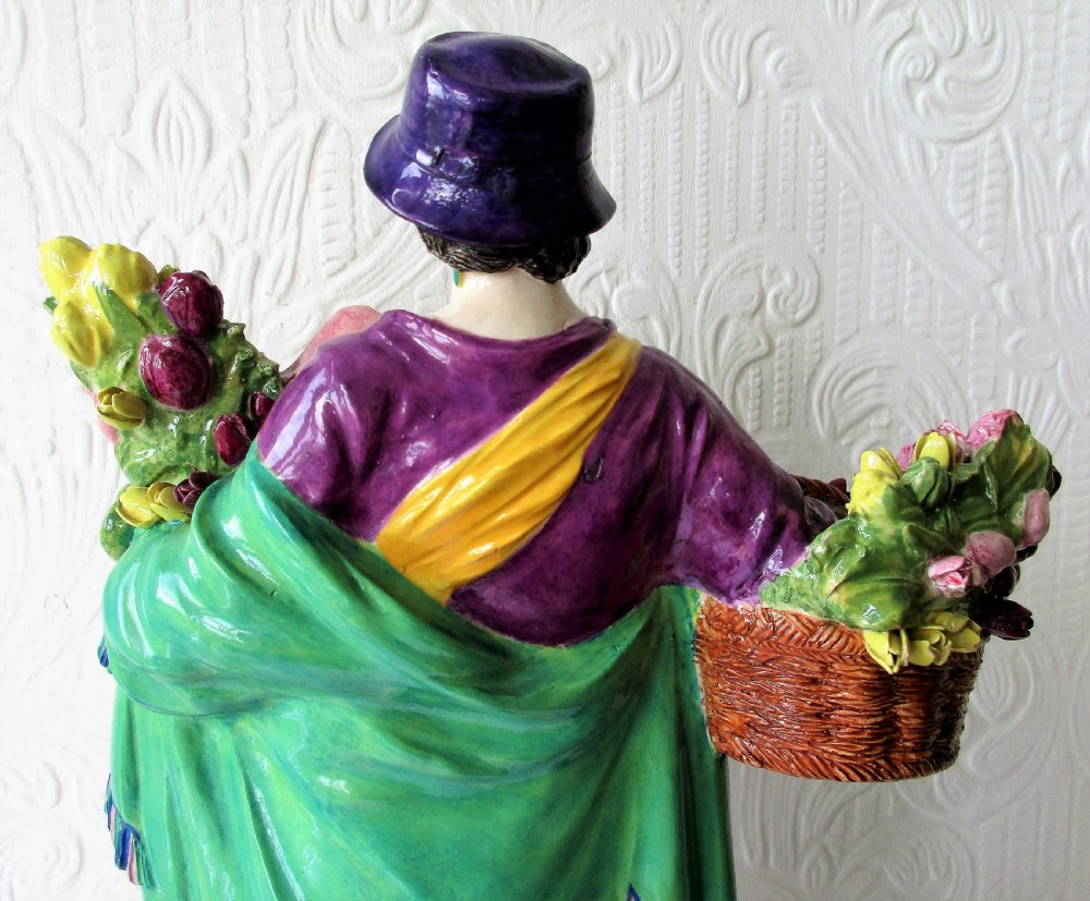 A polychrome glazed ceramic figurine depicting a woman wearing a purple jacket and blue hat, carrying a baby in a yellow sling and a basket of colourful flowers. View V