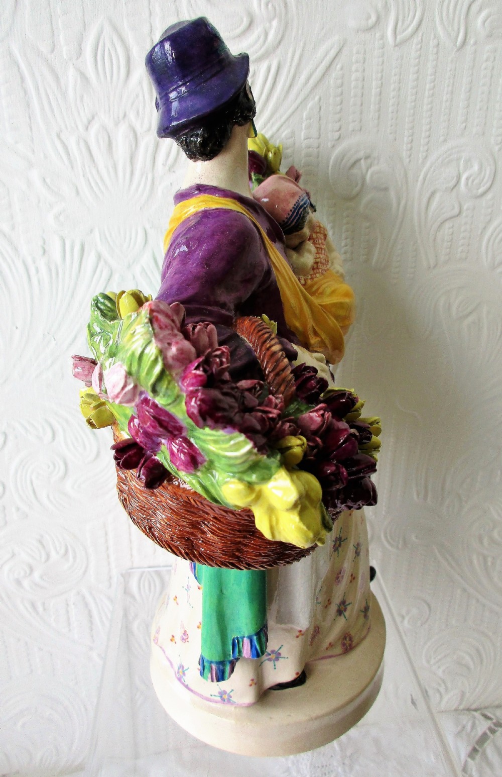 A polychrome glazed ceramic figurine depicting a woman wearing a purple jacket and blue hat, carrying a baby in a yellow sling and a basket of colourful flowers. View VI