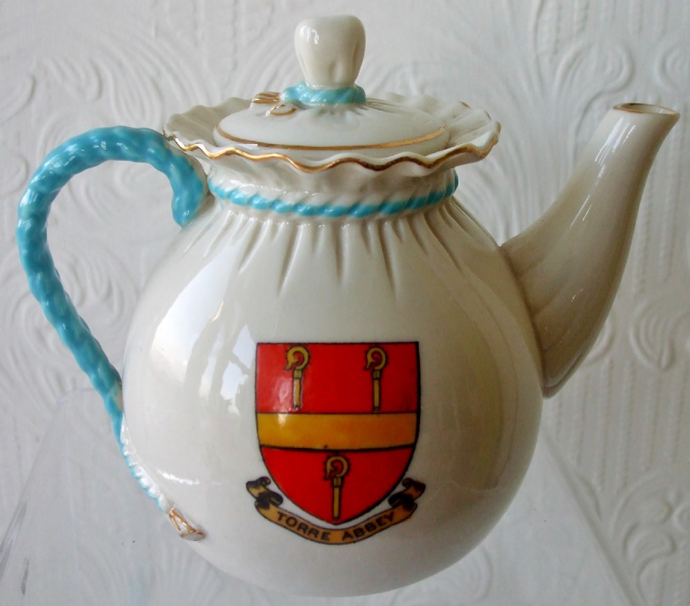 An antique white porcelain teapot shaped like a tied bag, featuring a blue twisted rope handle, a ruffled collar, and a coat of arms for Torre Abbey on the body.