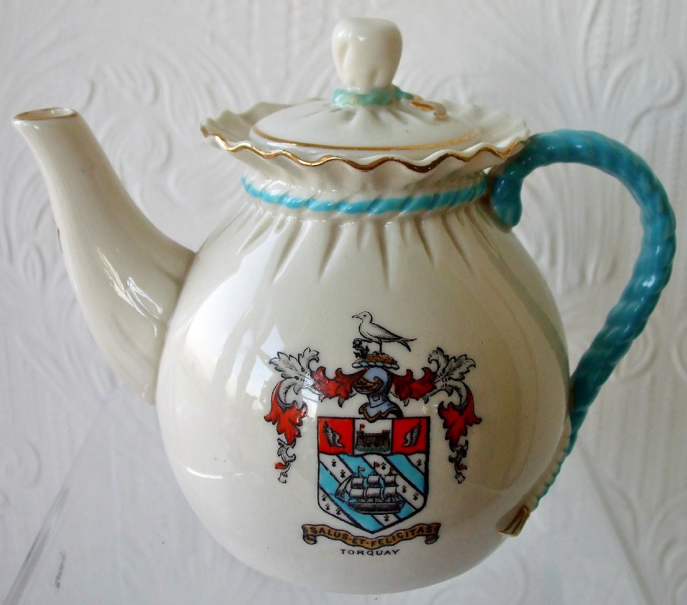 An antique white porcelain teapot shaped like a tied bag, featuring a blue twisted rope handle, a ruffled collar, and a coat of arms for Torre Abbey on the body. View III