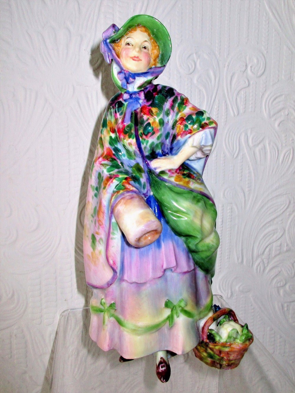 A hand-painted Royal Doulton bone china figurine depicting a female figure in a green bonnet and floral patterned cloak.