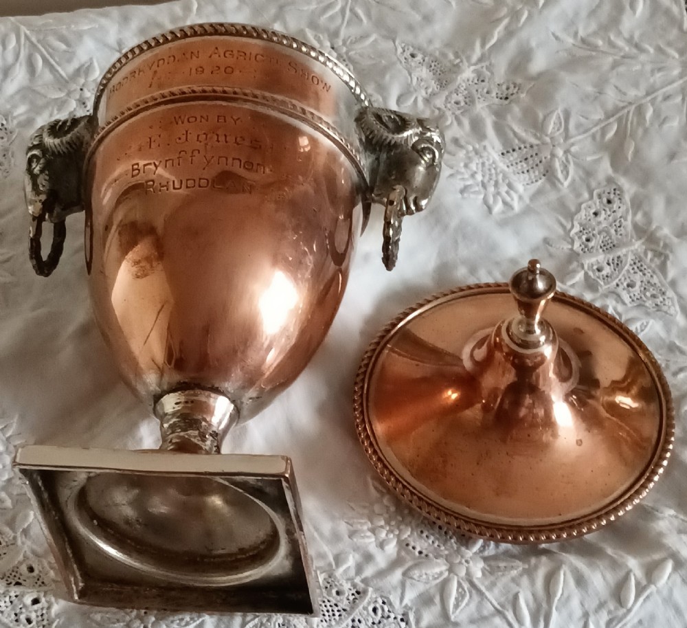 A miniature Georgian Old Sheffield Plate urn featuring a fitted lid with a finial, square base, engraved inscription, and rams head side handles. View V
