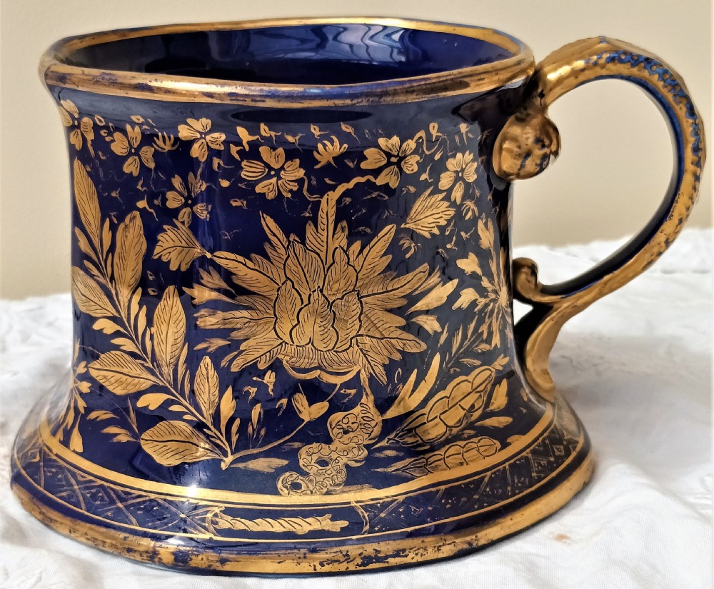 An antique cobalt blue ironstone mug featuring flared sides, a gilded scroll handle, and intricate gold floral motifs with a geometric border.
