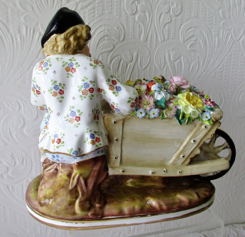 An English Victorian porcelain figurine depicting a seated male figure beside a wheelbarrow overflowing with colourful hand-crafted porcelain flowers. View III