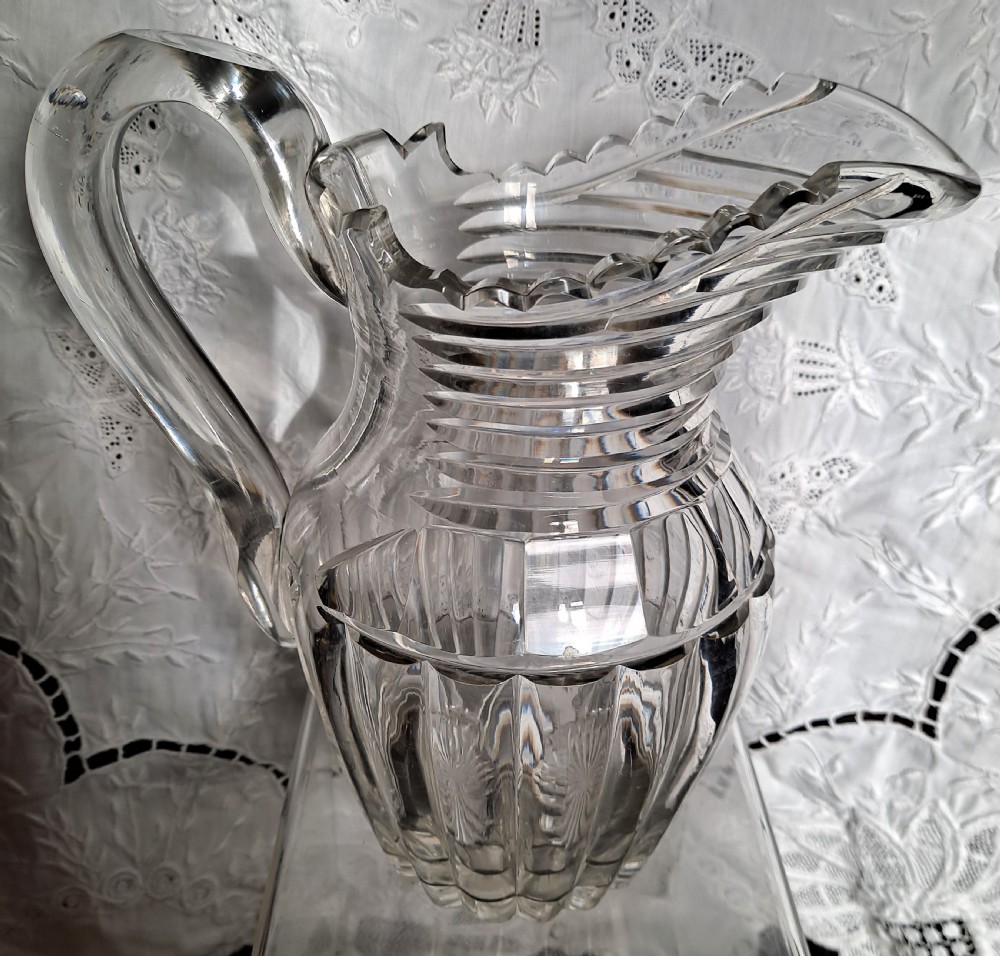A clear antique cut glass water jug featuring step-cut neck rings, deep vertical facet cutting, a scalloped rim, and a sturdy loop handle. View III