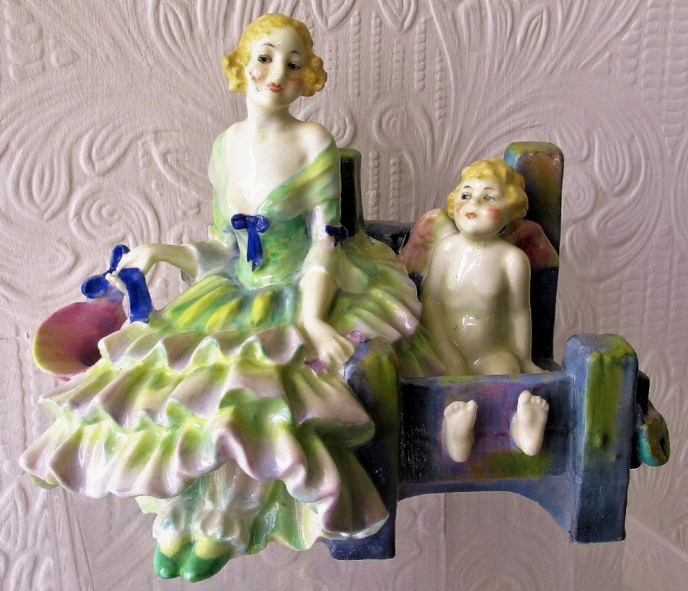An Art Deco Royal Doulton porcelain figurine depicting a lady in a green ruffled gown seated next to a cherub locked in wooden stocks.