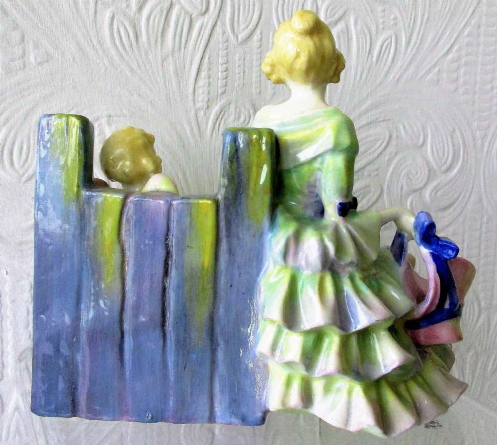 An Art Deco Royal Doulton porcelain figurine depicting a lady in a green ruffled gown seated next to a cherub locked in wooden stocks. View III
