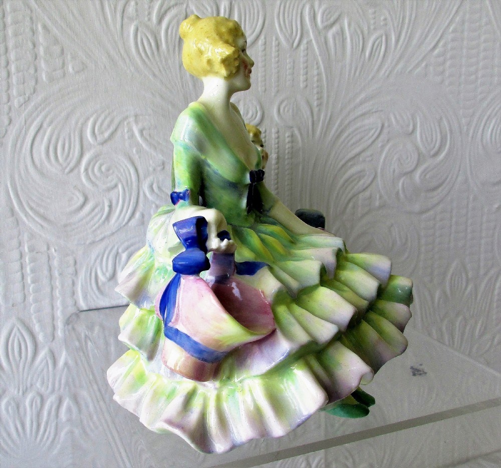 An Art Deco Royal Doulton porcelain figurine depicting a lady in a green ruffled gown seated next to a cherub locked in wooden stocks. View IV