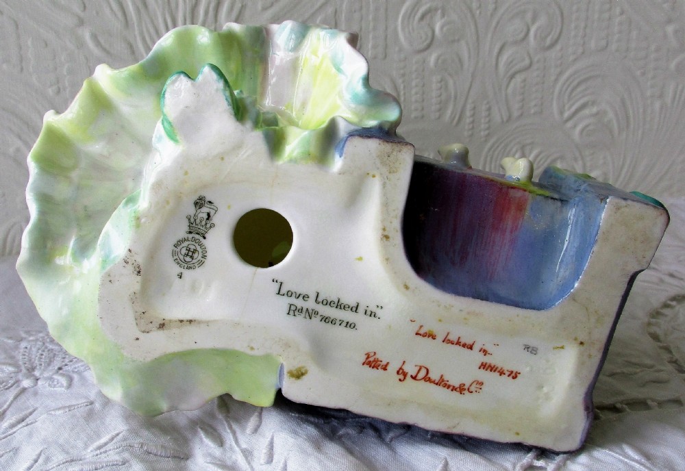 An Art Deco Royal Doulton porcelain figurine depicting a lady in a green ruffled gown seated next to a cherub locked in wooden stocks. View V
