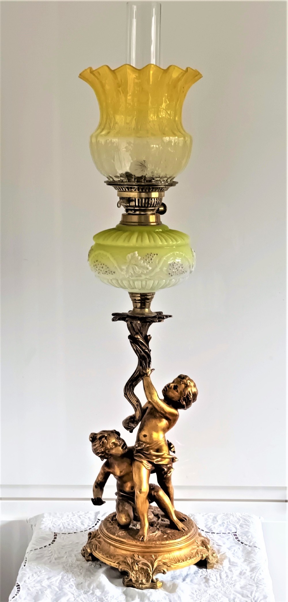 An antique Victorian oil lamp featuring a gilded metal base with sculpted cherubs, a green glass reservoir, and a yellow ruffled glass shade with chimney.