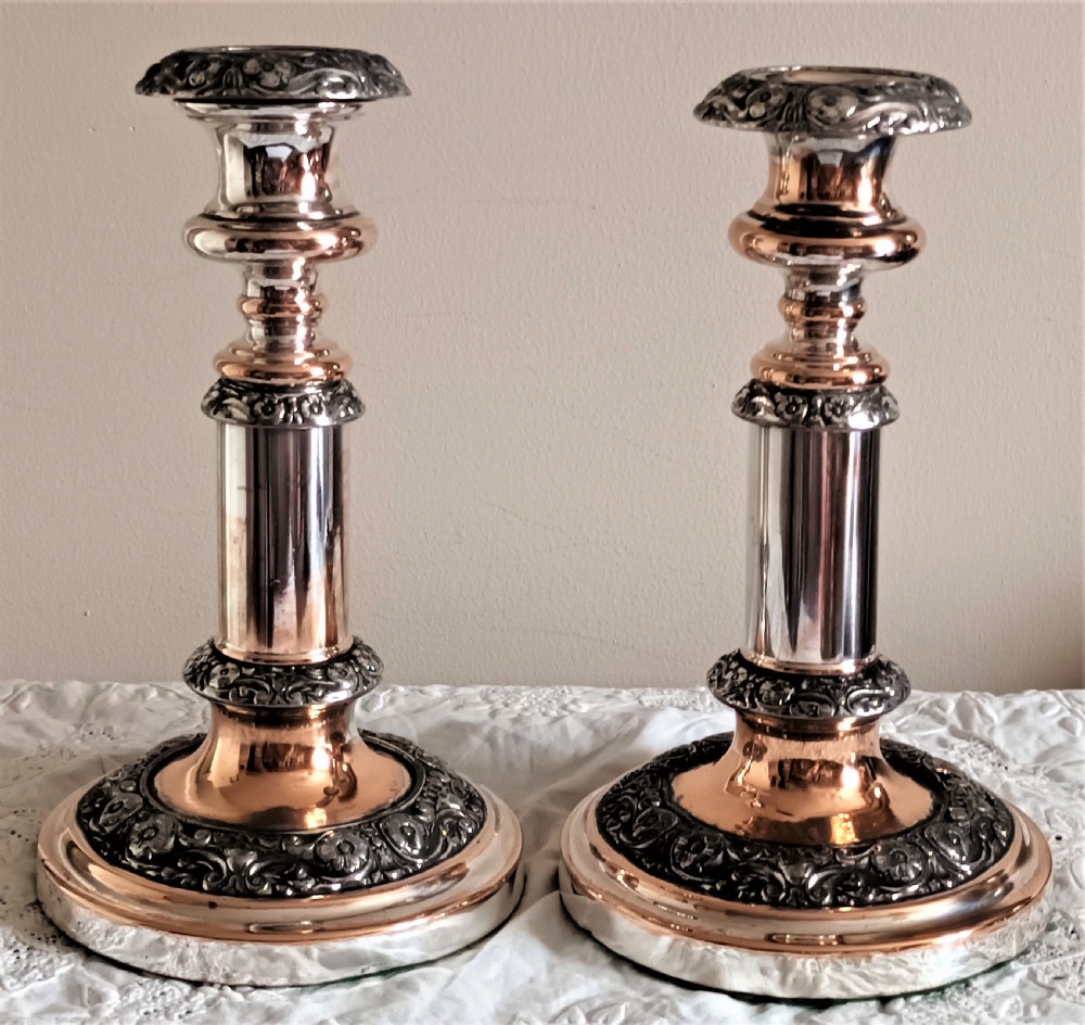 A pair of Georgian Old Sheffield Plate telescopic candlesticks featuring extendable silver columns showing copper undertones and ornate embossed borders.