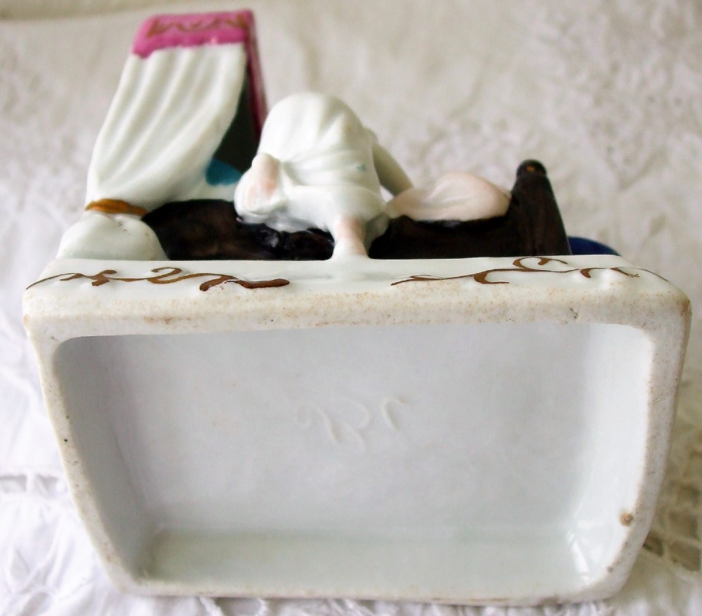 A small painted porcelain Victorian fairing depicting two figures wearing nightcaps in a canopy bed above an inscribed base. View V