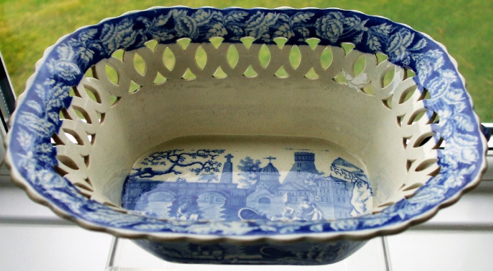 An oval Georgian blue and white ceramic chestnut basket featuring pierced lattice sides, a floral rim, and a central transfer-printed river landscape. View III