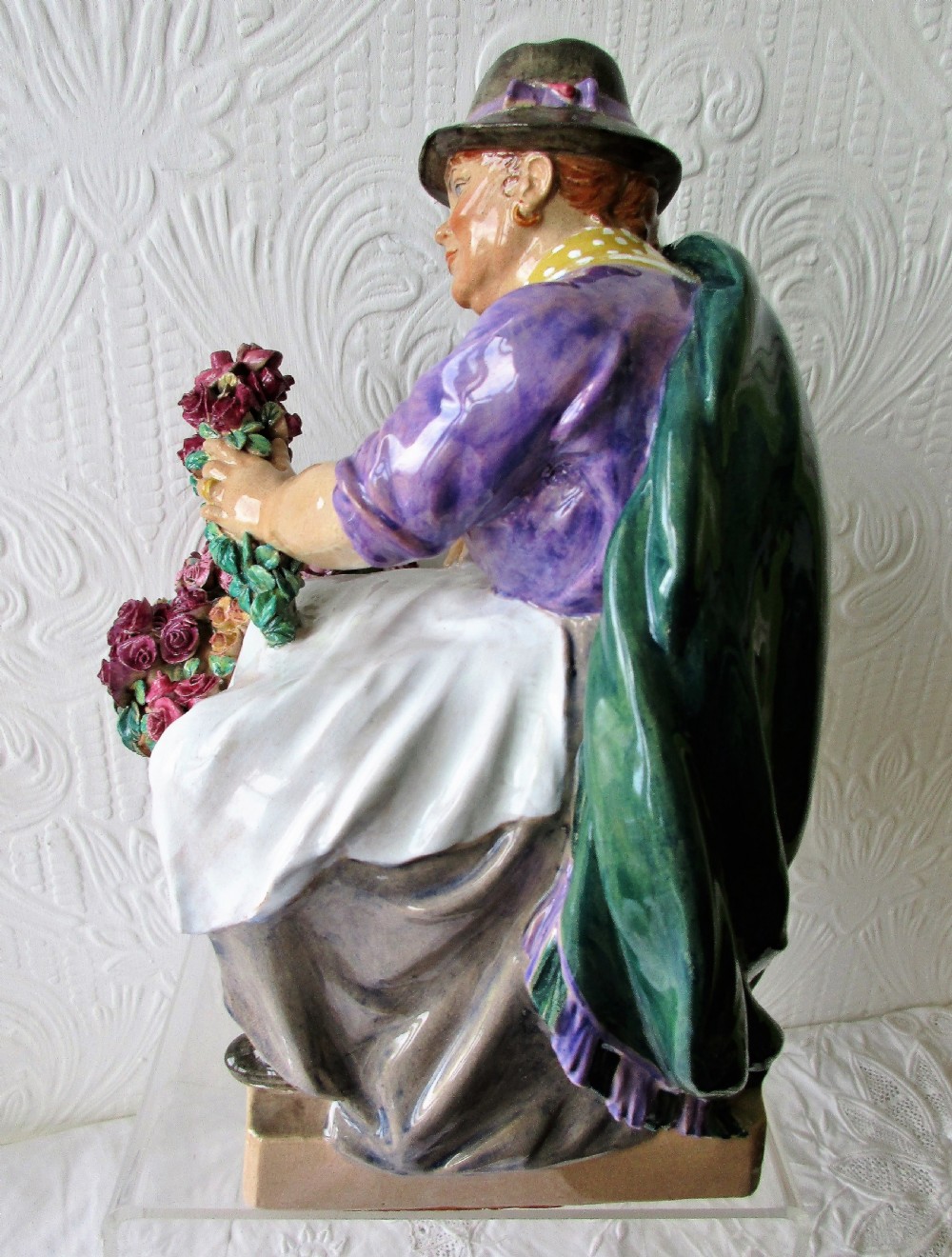 A polychrome glazed earthenware figurine depicting a seated woman wearing a green shawl and purple top holding a bouquet of red roses next to a basket full of flowers. View II