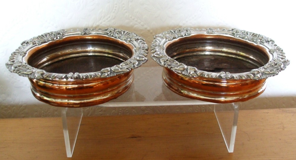 A pair of antique Georgian Old Sheffield Plate wine coasters featuring ornate relief borders and turned wood centres with exposed copper tone.