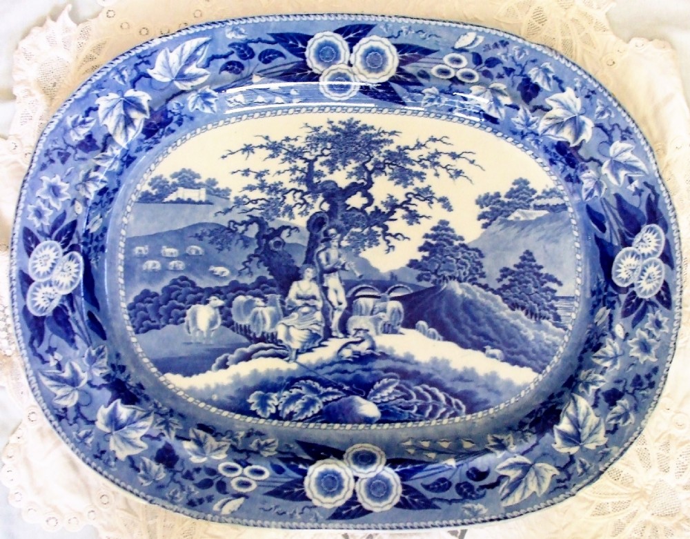 An oval antique Georgian earthenware serving platter featuring a blue and white pastoral scene with a piping shepherd surrounded by a floral border.