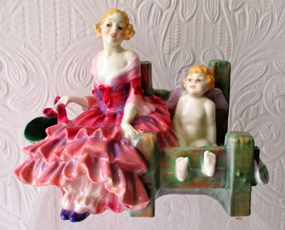 A hand-painted porcelain figurine depicting a woman in a ruffled pink dress seated alongside a small winged cupid trapped in green wooden stocks.