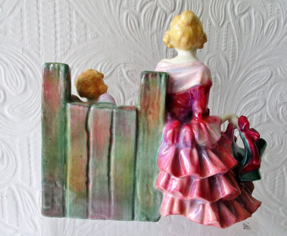 A hand-painted porcelain figurine depicting a woman in a ruffled pink dress seated alongside a small winged cupid trapped in green wooden stocks. View III