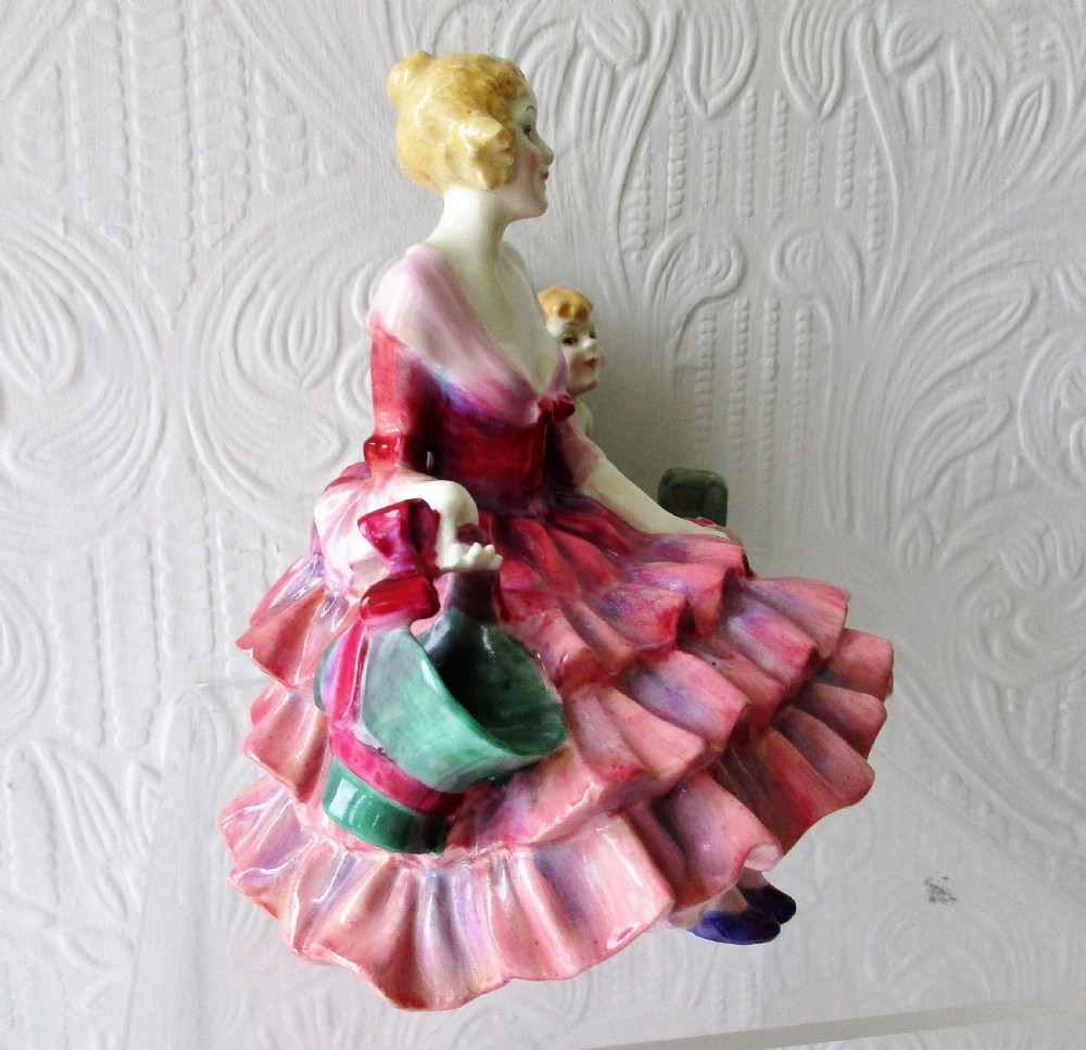 A hand-painted porcelain figurine depicting a woman in a ruffled pink dress seated alongside a small winged cupid trapped in green wooden stocks. View IV